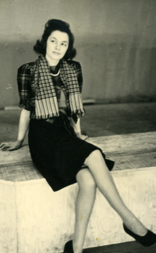 A teenage woman sitting on a stage in heels, a dress, and a scarf.