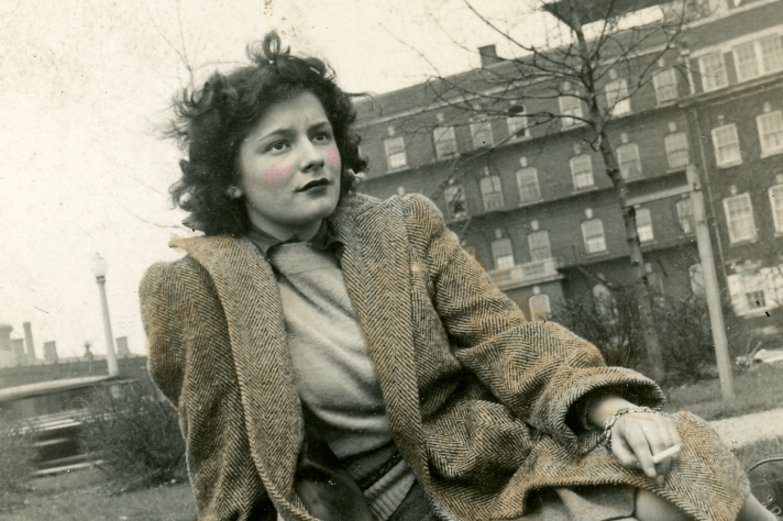 A black and white photo that has been colorized of a woman sitting in the grass in front of a brick building.
