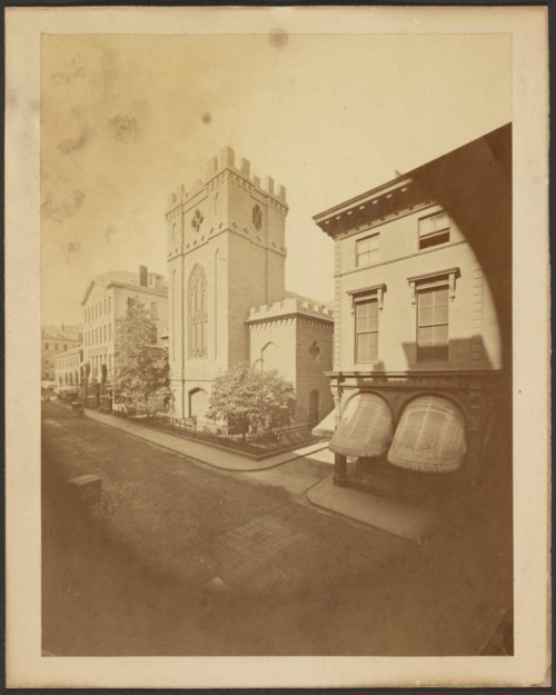 A sepia-toned photograph of a street, with a shop and a tall, crenellated church building.