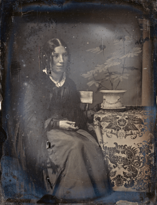 A black and white photograph of a seated woman. Next to her is a table with a patterned table cloth, which holds a vase with flowers.