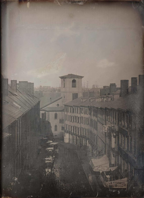 A black and white photograph of a city street. The photograph is taken from an aerial view, with buildings on either side and a procession taking place on the street below.