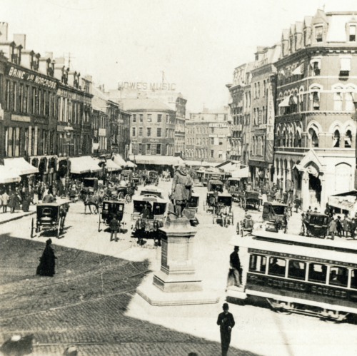 A black and white photograph of a bustling city square, filled with crowds and horse-drawn carriages.