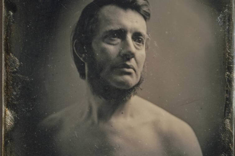 A shirtless man poses for a black and white photograph. He is middle aged with a beard.