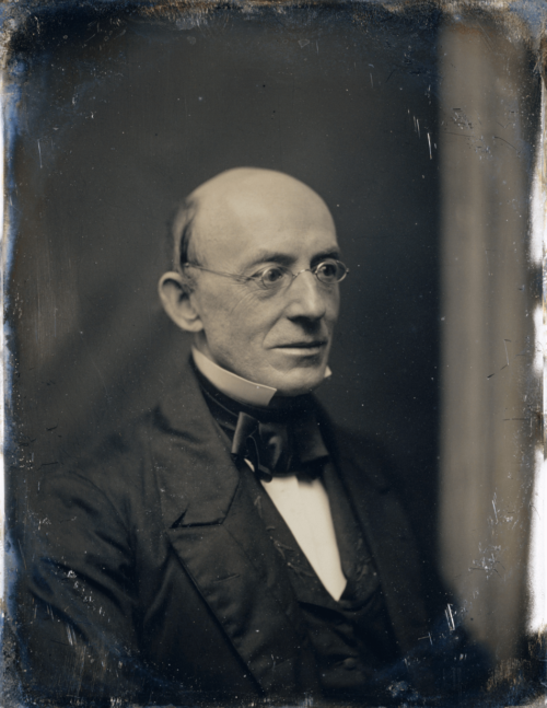 A man sits in a three quarters pose for his photograph. He wears a suit and glasses.