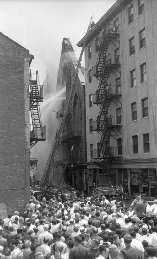 A large theater on fire. People stand below watching, as hoses spray water at the building.