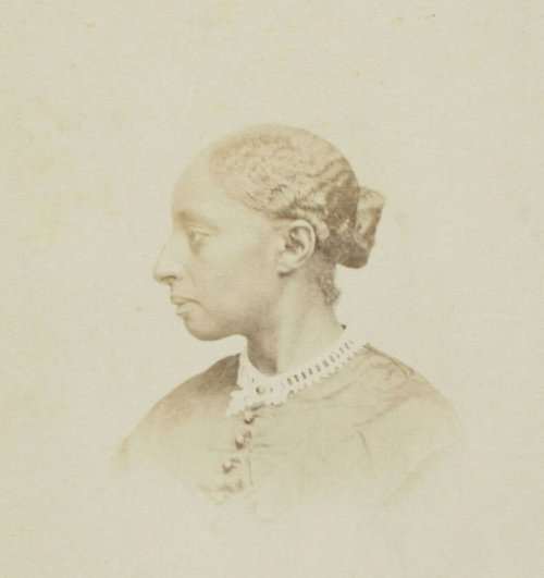 A photograph of a Black woman wearing a buttoned-up dress with her hair in a bun, looking sideways from the camera.