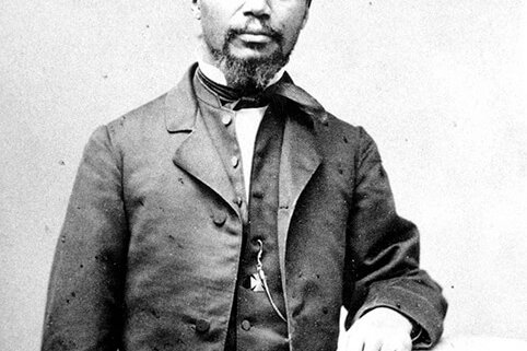 A Black man wearing a suit stands, facing the camera, with his arm rested on a column.