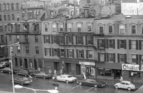 A black and white photograph of a city block, with lined four-story brick buildings.