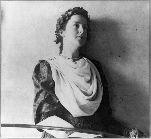 A black and white photograph of a woman leaning against a wall wearing a laurel around her head.