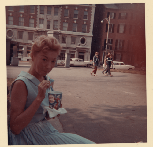 A photograph of a woman sitting on a park bench in a blue dress. She holds a cup with the straw in her mouth.