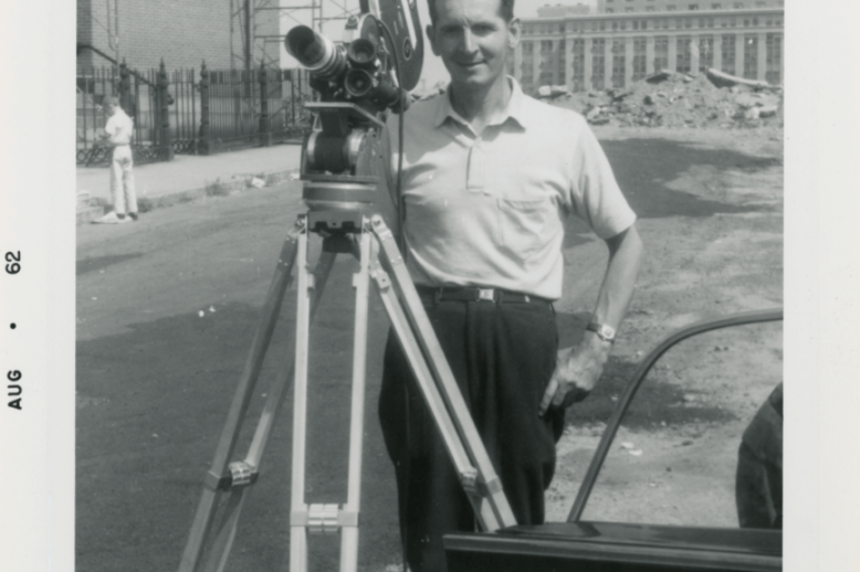 A black and white photograph of a man standing behind a movie camera with destroyed buildings in the background.