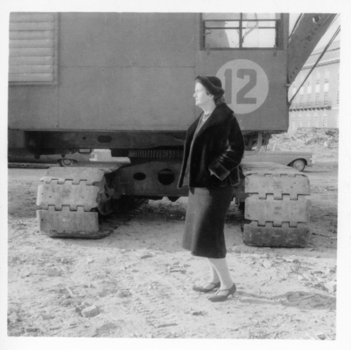 A black and white photograph of a woman wearing a dress, heels, and a jacket, standing in front of a crane.