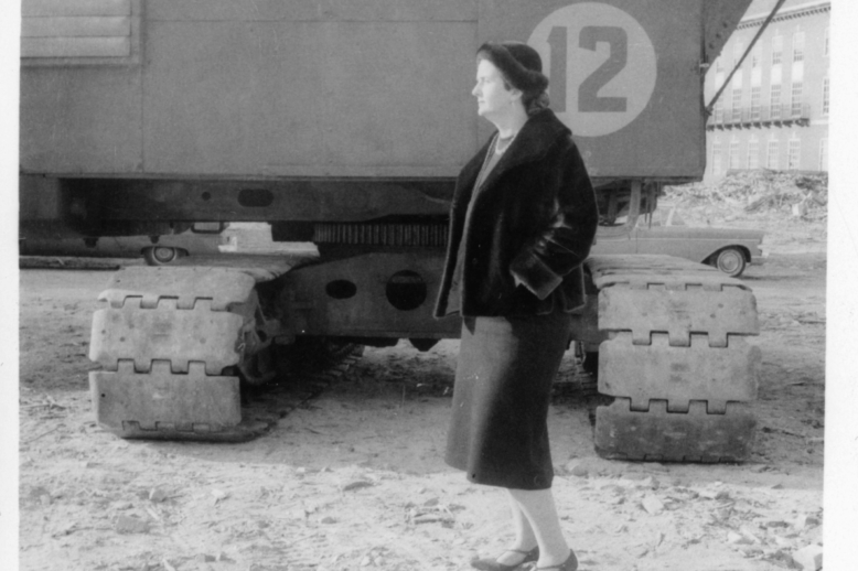Claudia Edgell A black and white photograph of a woman wearing a dress, heels, and a jacket, standing in front of a crane.