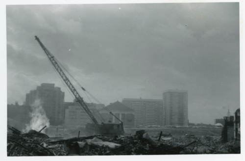 A black and white photograph of a crane and a wrecking ball. In the foreground, the rubble of destroyed buildings takes up most of the scene. In the background, some buildings remain standing.