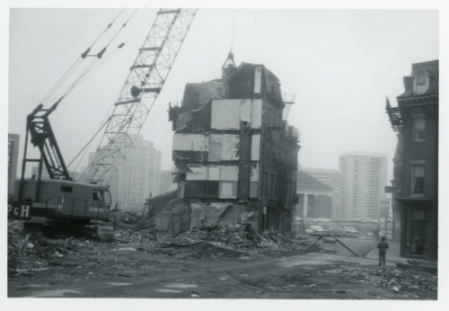 A black and white photograph of a crane and wrecking ball tearing down a residential building. Rubble and other destroyed buildings surround it.