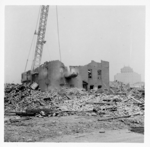 Black and white photograph of a building in the process of being demolished by a crane.