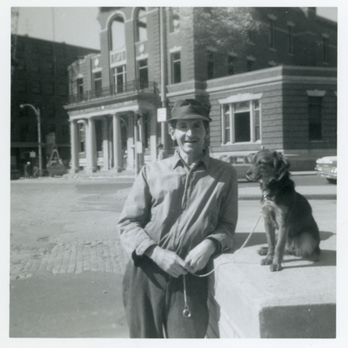 A black and white photograph of a man standing, holding a leash to a dog, who stands besides him.