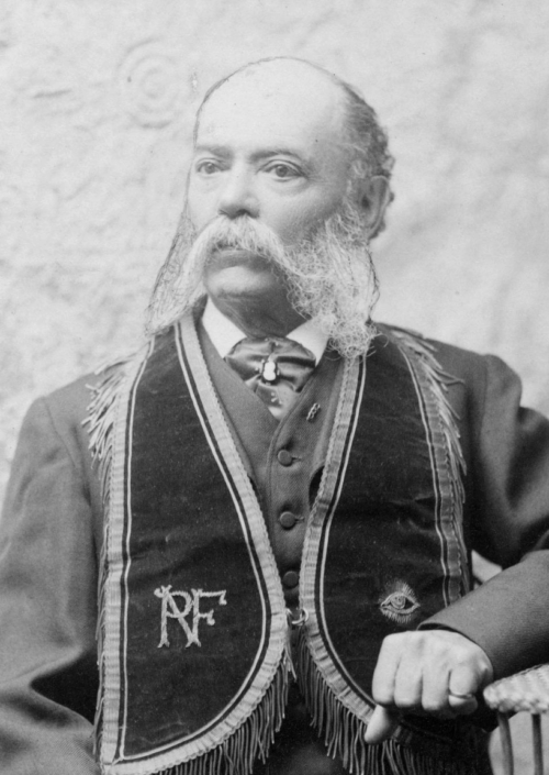 A black and white photograph of a man with a long white beard, sitting for a photograph in a suit.