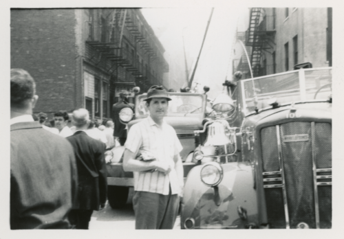 A black and white photograph of a crowd of people between buildings. Smoke and a fire truck are in the background.