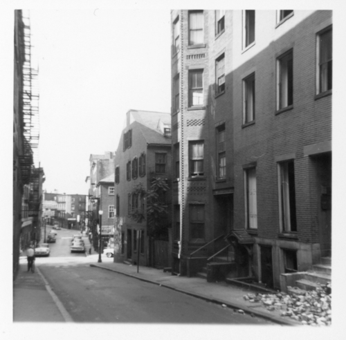 A black and white photograph of a city street, with four-story brick buildings side by side.