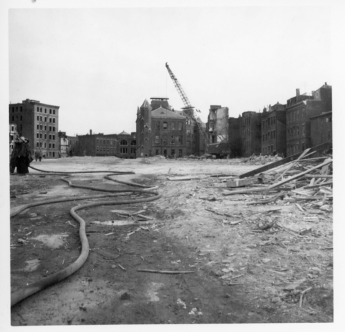A black and white photograph of an area cleared of buildings and filled with rubble. Some standing buildings and a crane stand in the background.