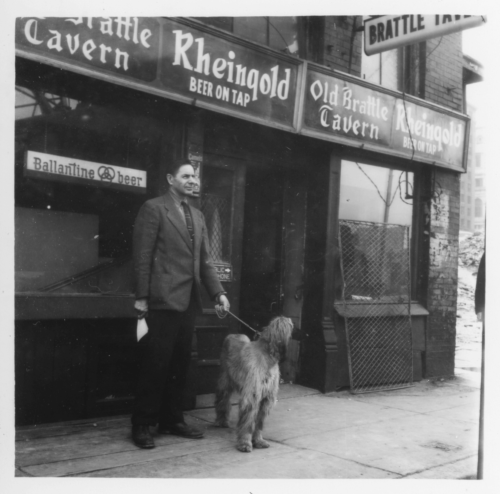 A black and white photograph of a man in a suit with a dog, standing in front of a tavern.
