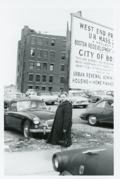 A black and white photograph of a woman standing in front of cars and a sign that reads "West End Project."