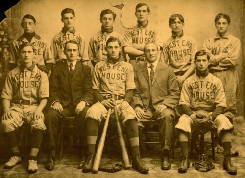 A sepia toned photograph of a group of young boys sitting for a team photograph. They wear jerseys that say "West End House" and one boy in the front holds baseball bats.