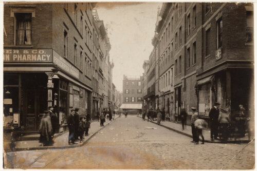 A sepia-toned photograph of a city street corner. People mill about in turn of the century clothing, and there is a pharmacy on the left.