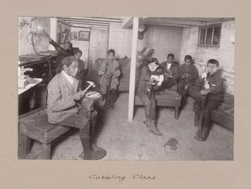 A group of White and Black young boys sit in a basement, repairing shoes.