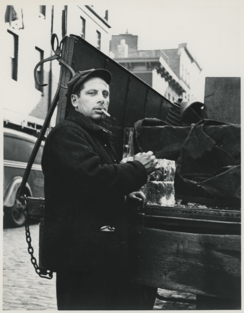 A black and white photograph of a man wearing a hat with a cigarette in his mouth. He is on a city street by the back of a truck, preparing to sell ice.