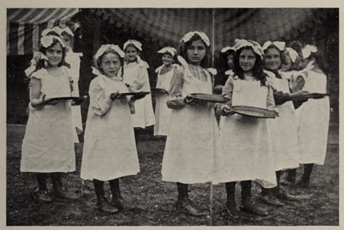 A black and white photograph of a group of young girls, wearing white frocks and carrying plates.