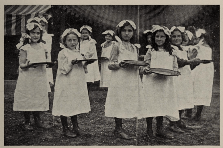 A black and white photograph of a group of young girls, wearing white frocks and carrying plates.