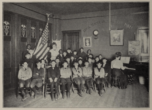 A black and white photograph of rows of boys sitting while a woman plays piano. They face the camera, with a prominent American flag at front.