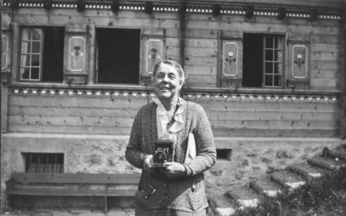A black and white photograph of a woman standing in front of a house, holding an old camera.