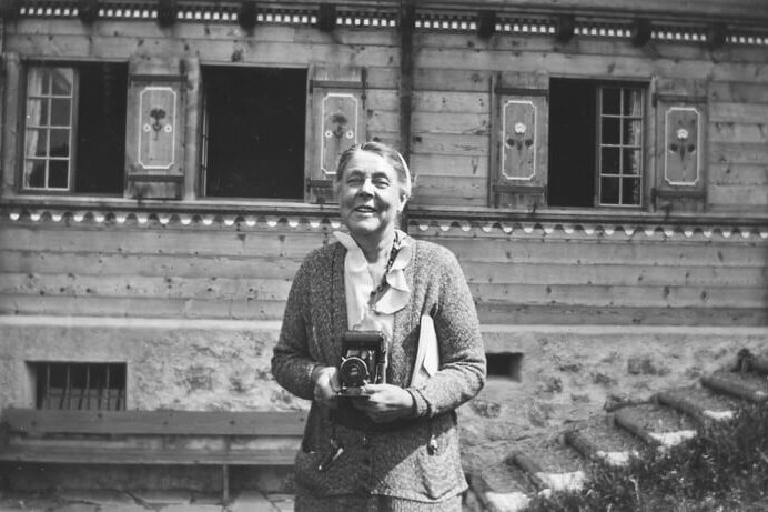 Helen Osborne Storrow A black and white photograph of a woman standing in front of a house, holding an old camera.