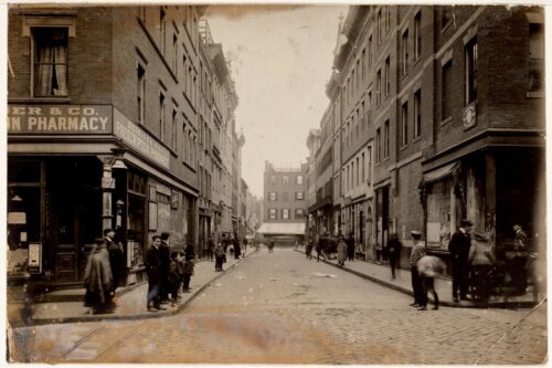 A sepia-toned view of Boston around 1910, showing a pharmacy and other businesses. The street is lined with people.