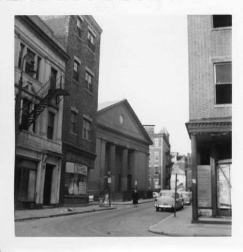 A black and white photograph of a city street. Between brick buildings stands a brick and stone neoclassical church.