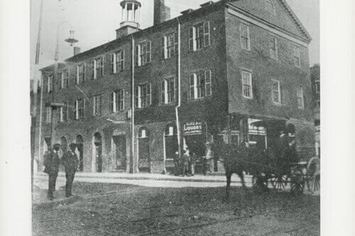 A black and white photograph of a three-story brick market building. People and horse-drawn carriages are in front of it.