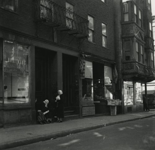 Black and white photograph of a street with three women waiting at a stoop.