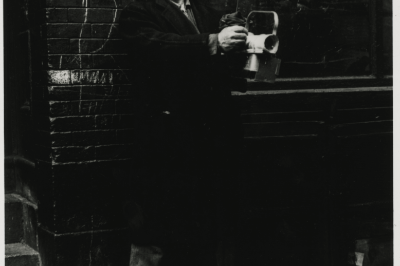 Tabachnik by Aarons A black and white photograph of a middle-aged man in a coat and hat, standing against a brick wall.