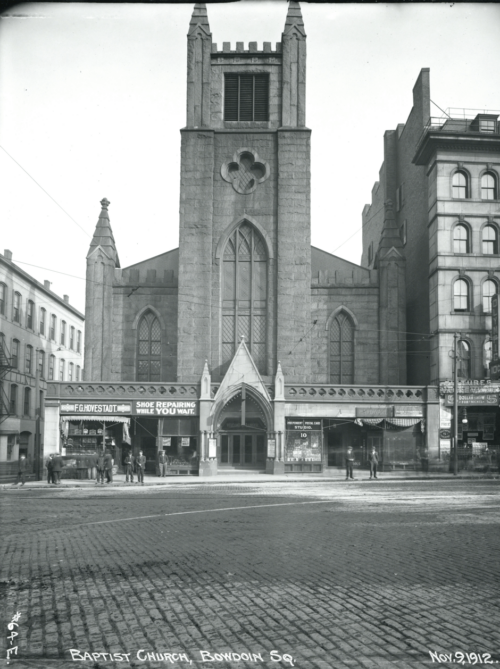 A black and white photograph of a neo-Gothic church in a city square, surrounded by brick and stone buildings.