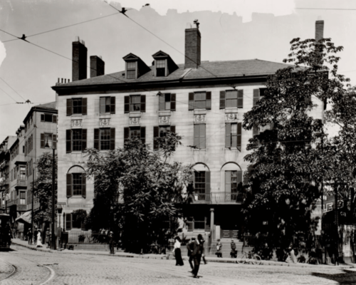 A black and white photograph of a large brick building at the end of a city square. People mill around in front of it.