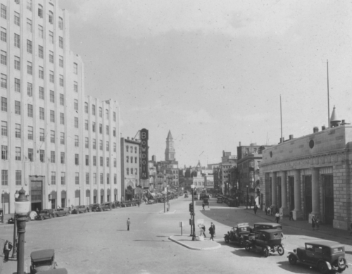 A black and white photograph of a street, with early 20th-century cars going down it. A short neoclassical firehouse is on the right, and a larger white building on the left.