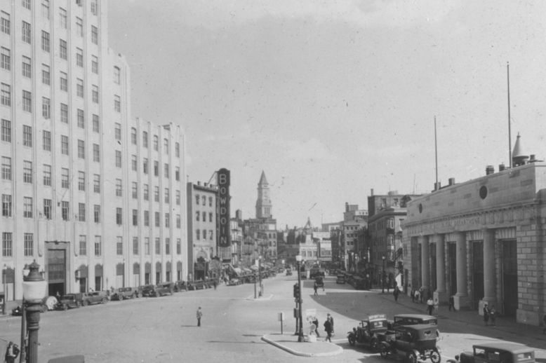 A black and white photograph of a street, with early 20th-century cars going down it. A short neoclassical firehouse is on the right, and a larger white building on the left.