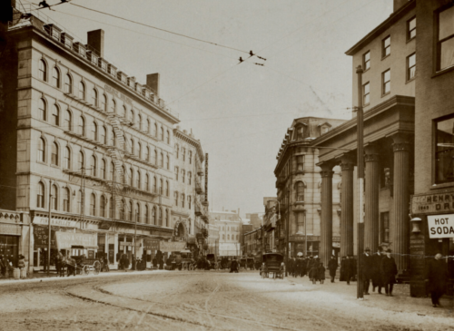 A sepia-toned photograph of an early 20th-century city square. The street is cobblestone, people in suits mill about, and the buildings are about 6 stories tall.
