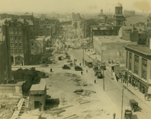 A sepia-toned image of construction on an early 20th-century street. Parts of the street are upturned, and on the sides are brick buildings and a church.