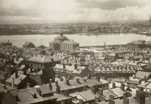 A black and white photograph from above showing rows of brick buildings. In the background there is a river with a large granite building next to it, the Charles Street Jail.
