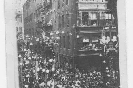 A black and white photograph of a city block, lined with 4-5 story brick buildings, with crowds of people in the street.