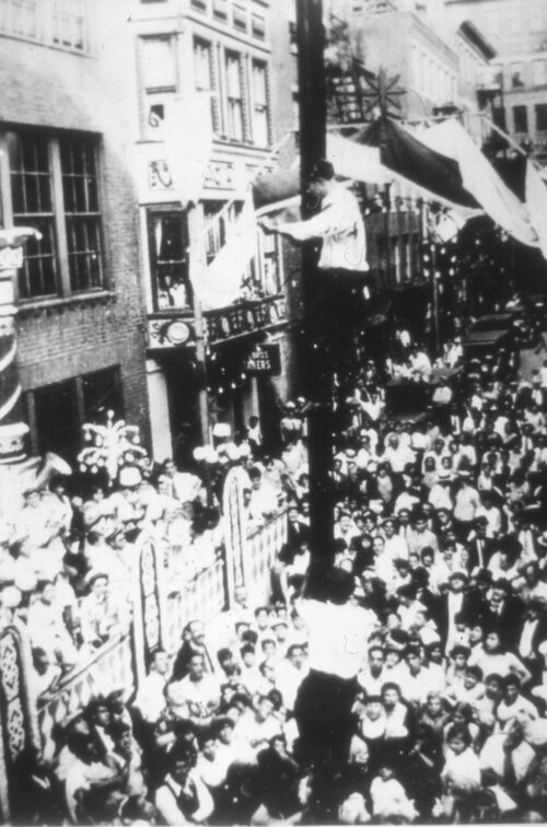 A black and white photograph of a crowd of people on a city street. In the center, two men attempt to climb a slippery pole.